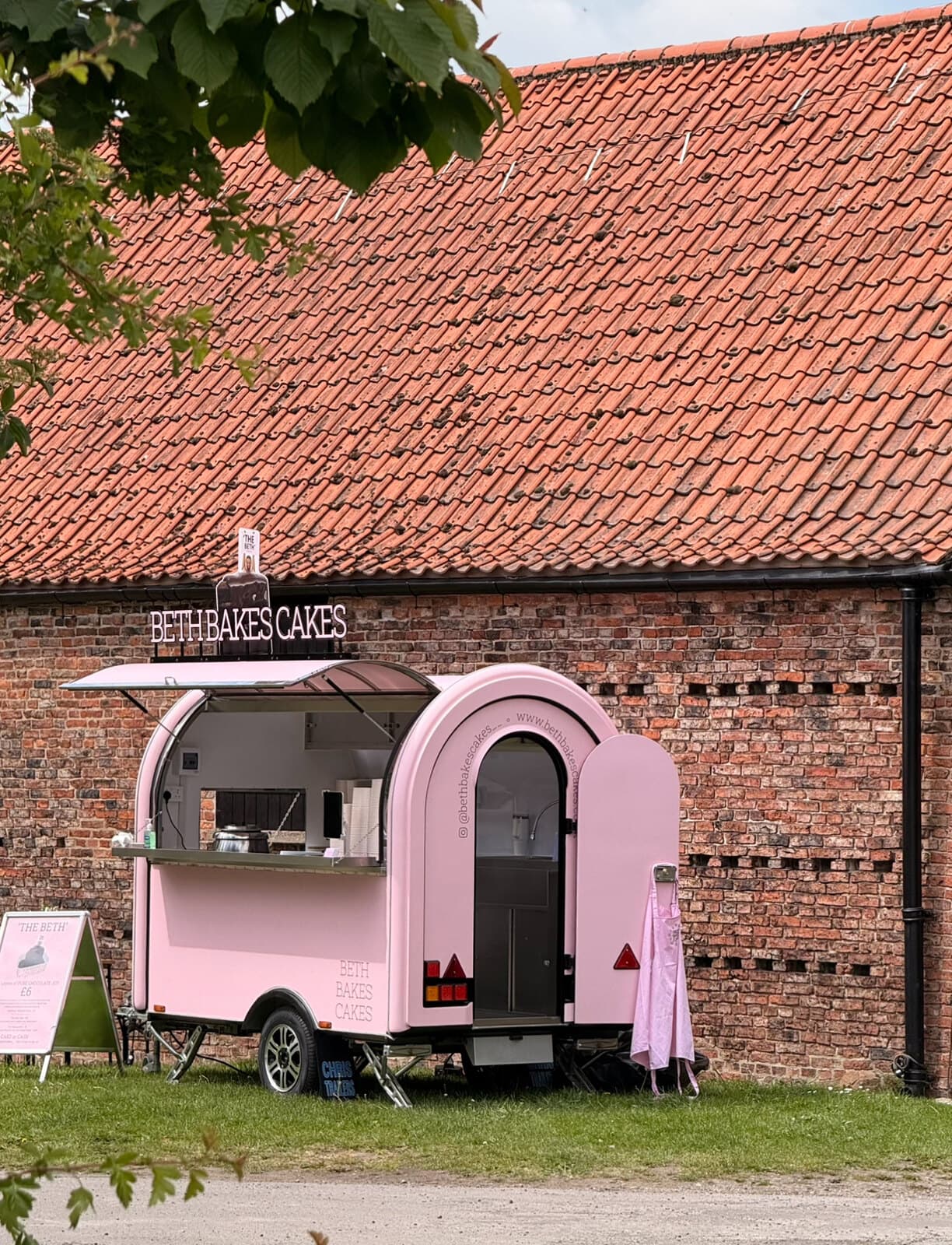 'The Beth', a pink trailer bar with a BETH BAKES CAKES sign, parked in front of a brick barn